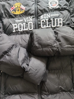 Svart pufferjacka Vinson Polo Club - Svart pufferjacka från Vinson Polo Club med broderade loggor och text på bröstet. Jackan har hög krage, dragkedja framtill och quiltad design. Perfekt för kalla dagar och ger en sportig look.