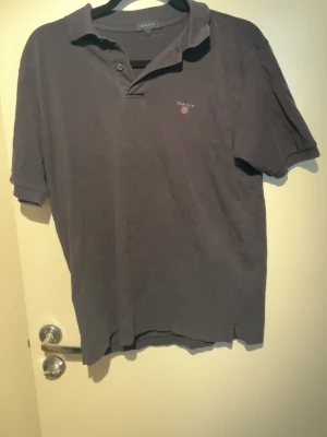 Mörkgrå pikétröja från Gant - Klassisk mörkgrå pikétröja från Gant med korta ärmar och liten logga på bröstet. Tröjan har krage och knappar vid halsen. Perfekt till jeans eller shorts för en avslappnad stil.