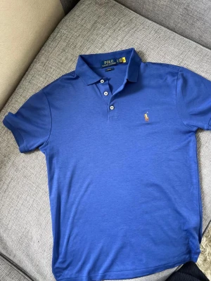 Blå pikétröja från Polo Ralph Lauren - Klassisk blå pikétröja från Polo Ralph Lauren med korta ärmar och krage. Tröjan har tre knappar framtill och den ikoniska broderade loggan på bröstet. Perfekt till jeans eller shorts för en clean och stilren look.