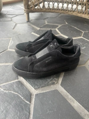 Svarta Arigato laceless i strl 45 - Stilrena svarta sneakers från Axel Arigato i mocka med diskret logga på sidan. Skorna har en minimalistisk design utan synliga snören och en svart gummisula. Perfekta för dig som gillar en clean och modern look.