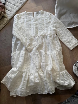 Vit långärmad klänning från Zara - Superfin vit klänning från Zara med lång ärm och volangdetaljer. Klänningen har en strukturerad yta och är tillverkad i ett lätt material som passar perfekt till varmare dagar. Den har en avslappnad passform och rund halsringning.