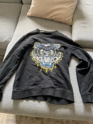 Kenzo Hoodie - Cool svart sweatshirt från Kenzo med ikoniskt tigerbroderi i blått, gult och vitt på bröstet. Rund halsringning och ribbade muddar. Perfekt för dig som vill sticka ut med en streetstyle-look.