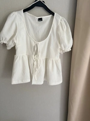 Vit blus med puffärm och knyt - Vit blus från Gina Tricot med korta puffärmar och knytning framtill. Tillverkad i ett lätt, strukturerat bomullstyg med volang nedtill och djup rundad urringning. Perfekt för en söt och trendig look.