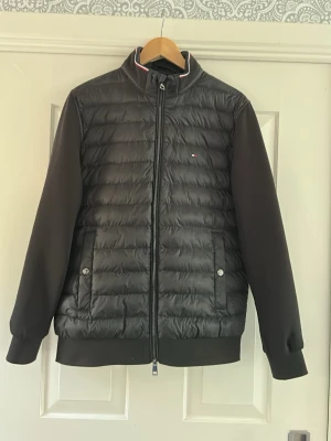 Svart pufferjacka från Tommy Hilfiger - Snygg svart pufferjacka från Tommy Hilfiger med quiltad front och släta ärmar. Jackan har dragkedja, ståkrage med diskret rödvit detalj och två fickor med tryckknappar. Liten logga på bröstet och ribbade muddar vid ärmslut och nederkant.