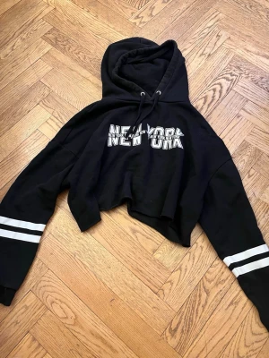 Svart cropped hoodie  - Svart cropped hoodie med vit text 'New York' på bröstet och dubbla vita ränder på ärmarna. Huva med dragsko och långa ärmar. Perfekt för en avslappnad streetstyle-look. Passar S-M-L