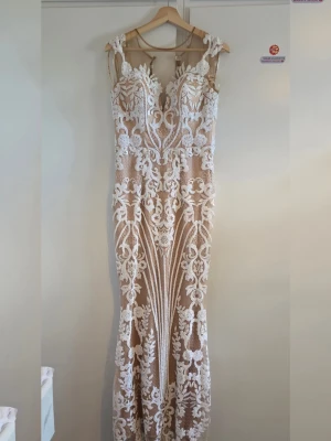 Lång spetsklänning med blommönster - Säljer nu denna fantastiska långklänning med invecklad benvit spets över en nude/champagnefärgad bas. Klänningen har en smickrande sjöjungfru-siluett (fit and flare) som framhäver figuren vackert. Storlek 38 och använd en gång bara. Sista bilden visar defecten som är längst ner. 