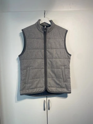 Grå dunväst från Berkeley - Snygg grå dunväst från Berkeley med ståkrage och dragkedja framtill. Västen har två fickor med lock och quiltad design som ger en stilren look. Perfekt att slänga över en hoodie eller tröja för extra värme.