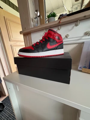 Air Jordan 1 mid  Svart/Röd - Snygga Air Jordan 1 mid sneakers i svart och rött läder med klassisk röd Swoosh och vita sulor. Skorna har röda skosnören och ikoniska Jordan Wings-loggan på hälen. Perfekta för dig som gillar streetwear och vill sticka ut. Skorna är storlek 37,5 och har andvänts runt 2-3 gånger. Dem kommer inte i original skoboxen pga att jag har tappat bort den. Skriv för mer bilder