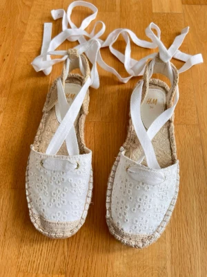 Vita espadrilles-sandaler med knytband - Säljer ett par vita espadrilles-sandaler från H&M med broderade blommor och långa knytband som lindas runt ankeln. Ovandelen är i tyg med broderidetaljer och sulan är i flätad jute. Perfekta för en somrig och avslappnad stil.