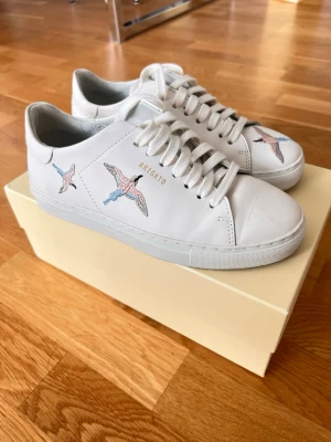 Vita sneakers från Axel Arigato - Snygga vita sneakers från Axel Arigato i läder med broderade fåglar i blått och rosa på sidan och tån. Klassisk låg modell med vita skosnören och diskret guldfärgad logga på sidan. Perfekta för dig som vill ha en clean look med en unik detalj. Storlek: 36. Säljer för att de visade sig vara för små i storleken för mig, använt dem 2 gånger ute. Köptes för ca 2300kr 