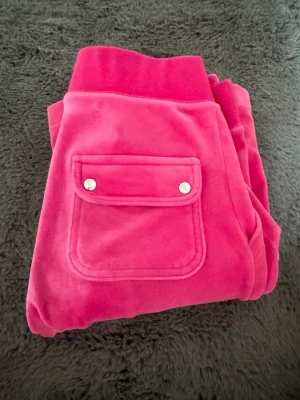 Juciy byxor - Snygga rosa mjukisbyxor i mjuk velour med bred resår i midjan och praktiska fickor, varav en med lock och silvriga knappar. Perfekta för en chill och trendig look. Materialet är lent och stretchigt för extra komfort.