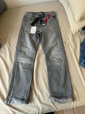 Grå raka jeans från replay Thad - Snygga grå jeans från replay med raka ben och klassisk femficksdesign. Jeansen har en diskret tvättad look och svarta detaljer vid midjan. Perfekta för en avslappnad och trendig stil.