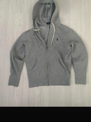 Grå hoodie från Polo Ralph Lauren - Grå zip-hoodie från Polo Ralph Lauren med klassisk huva och vita dragsnören. Broderad blå logga på bröstet och två fickor framtill. Mjuk bomullsblandning som är skön att ha på sig. Perfekt för en avslappnad stil.