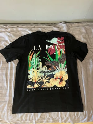 Svart LA PAZ t-shirt med tryck - Svart t-shirt från Jack&Jones med färgglatt tryck av växter och texten 'LA PAZ' på ryggen. Framtill finns en liten logga och text. Klassisk passform och rund hals. Perfekt för dig som gillar statement-plagg och grafiska motiv.