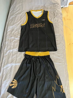 Svart Raptors basketset med gula detaljer - Snyggt basketset med linne och shorts i svart med gula detaljer och Raptors-tryck. Linnet har gul kant vid hals och ärmar, och shortsen har elastisk midja samt tryck med basketboll och blixt. Perfekt för basketträning eller streetstyle.