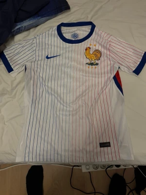 Frankrike fotbollströja Nike - Säljer en vit Frankrike fotbollströja från Nike med tunna blå och röda ränder. Tröjan har korta ärmar, blå krage och är prydd med det klassiska tupp-emblemet och två stjärnor på bröstet. Perfekt för dig som älskar fotboll och vill visa ditt stöd för landslaget.