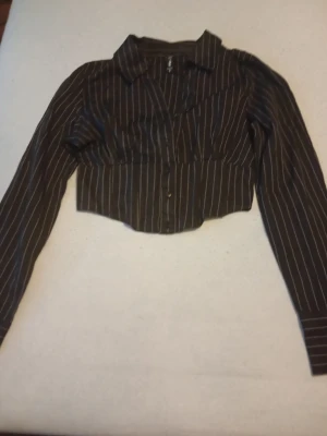 Svart croppad skjorta med ränder - Snygg svart croppad skjorta från H&M med vita vertikala ränder. Skjortan har klassisk krage, knäppning framtill och långa ärmar. Perfekt för dig som vill ha en trendig och stilren look.