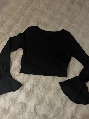Svart croppad topp med trumpetärm - Svart croppad topp med långa trumpetärmar och rund halsringning. Toppens stilrena design gör den enkel att matcha med olika outfits. Tillverkad i mjukt material som känns skönt mot huden.