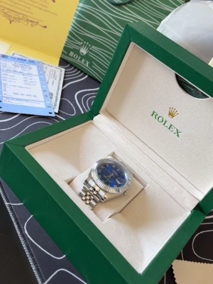 Clean klocka med grön klockbox med tillbehör - Säljer en exklusiv klocka med klassisk design. Boxen har en lyxig känsla och kommer med tillhörande certifikat och påse. Perfekt för dig som vill förvara eller presentera din klocka på ett snyggt sätt. Med en speciell känsla av den silvriga och blåa delen med detta mästerverk, kommer även med påse och papper 