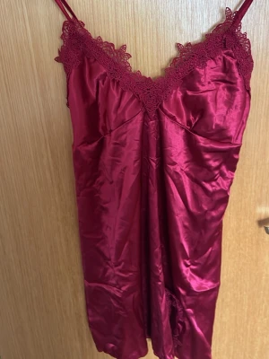 Vinrött satin nattlinne med spets - Snyggt vinrött nattlinne i glansigt satinmaterial med tunna axelband och dekorativ spets vid urringning och nederkant. Perfekt för en lyxig känsla hemma. Figurnära passform och mjukt tyg som känns skönt mot huden.