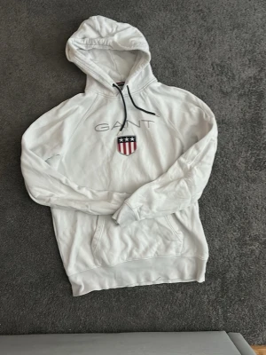 Vit hoodie från GANT med logga i mitten. -  Stocholm Deluxe design Vit hoodie från GANT med broderad logga och amerikanskt sköldmärke på bröstet. Klassisk känguruficka framtill och justerbar huva med marinblå snören. Perfekt för en avslappnad och sportig stil, fungerar även bra till Stocholm stilen. Storlek L/M