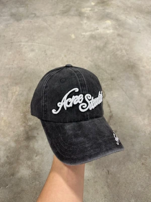 Svart/grå  keps från Acne Studios - Svart/grå keps från Acne Studios med vit broderad logga framtill och på skärmen. Klassisk böjd skärm och justerbar passform. Tillverkad i mjuk bomull med en tvättad look som ger en avslappnad vibe.