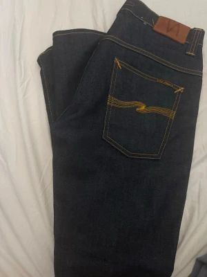Mörkblå jeans från Nudie Jeans - W29 L32Säljer ett par mörkblå jeans från Nudie Jeans med klassisk gul kontrastsöm och vågig brodyr på bakfickorna. Jeansen har rak passform och är tillverkade i kraftig denim. Perfekta för dig som gillar stilrena och tidlösa jeans.modell tape Ted 