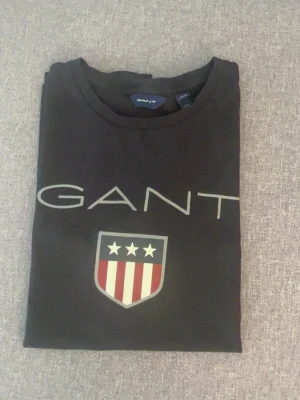 Gant T-Shirt - Svart t-shirt från Gant med stor logga och sköld med stjärnor och ränder på bröstet. Klassisk rund hals och korta ärmar. Perfekt för sommaren som kommer. 