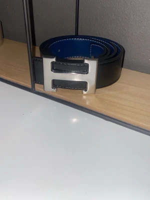 svart/mörkblått bälte - Snyggt svart bälte som man kan vända till mörkblått med ett H i silver som logga.