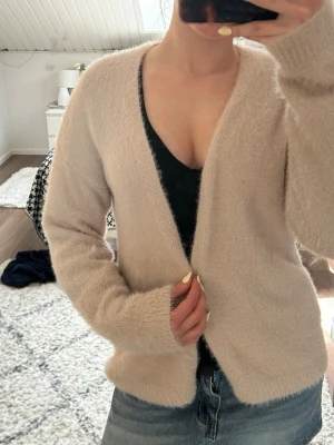 Beige fluffig kofta utan knappar - Mysig beige kofta i fluffigt material med lång ärm och öppen framtill utan knappar. Perfekt att slänga över ett linne eller t-shirt för en avslappnad look. Passar till både jeans och kjol för en enkel och trendig stil.