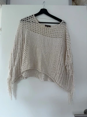 Beige stickad poncho med fransar - Säljer en beige stickad poncho med fransar längs kanterna. Ponchon har ett luftigt, grovmaskigt mönster upptill och längs ärmarna, samt tätare stickning framtill. Perfekt att slänga över en topp för en chill look.
