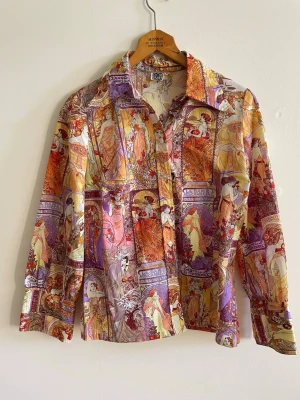 Bon Bee vintage skjorta i Art Nouveau-stil – Unikt statementplagg storlek 42 - Unik vintageinspirerad skjorta från Bon Bee med fantastiskt Art Nouveau / Alphonse Mucha-inspirerat mönster. Ett riktigt statement-plagg med konstnärliga detaljer och vackra toner i orange, lila, guld och creme.  Detta är ett riktigt fynd för den som älskar vintage och ovanliga plagg. Mönstret ger en exklusiv retro- och editorial-känsla och passar perfekt till kjol, byxor eller lager-på-lager styling.  Storlek: 42 Skick: Bra vintageskick  Perfekt för: • vintage-estetik • retroälskare • konstnärl