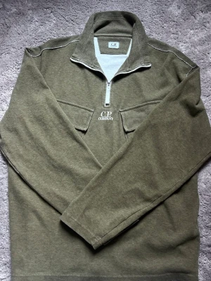 Olivgrön halfzip från C.P. Company - Olivgrön långärmad tröja från C.P. Company med halv dragkedja och hög krage. Tröjan har två bröstfickor och vit logga broderad framtill. (Köptes för 3000kr).