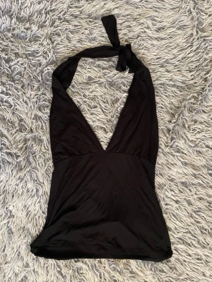 Svart halterneck topp med knyt - Snygg svart halterneck topp med djup v-ringning och knyt i nacken. Toppens material har en lätt glansig finish som ger en cool look. Perfekt att styla med jeans eller kjol för en trendig outfit. Passar även någon i XS, XSS, S då man kan knyta!