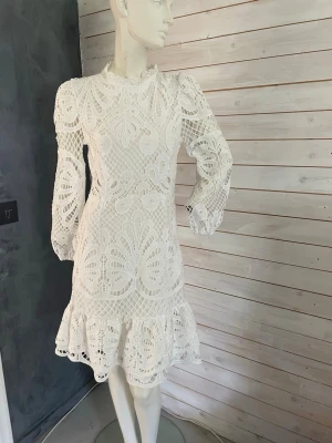 Vit spetsklänning med volang - Superfin vit klänning i spets med lång ärm och volang nertill. Klänningen har hög hals, markerad midja och dragkedja i ryggen. Detaljerad spets med blommigt mönster ger en romantisk känsla. Perfekt för sommarens tillfällen.