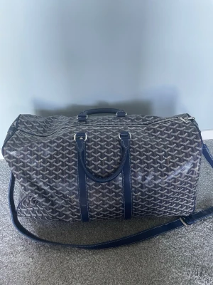 Blå weekendbag från H&M - Snygg och rymlig weekendbag i blått med klassiskt mönster. Väskan har både handtag och justerbar axelrem samt silverfärgade detaljer. Perfekt för resor eller gymmet, med dragkedja upptill och stilren design.