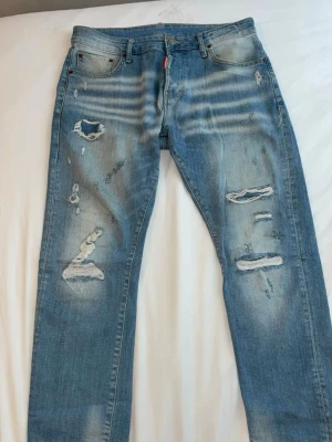 Ljusblå slitna jeans från DSQUARED2 - Säljer ett par ljusblå jeans från DSQUARED2 med coola slitningar och hål på framsidan. Jeansen har klassisk femficksdesign, raka ben och tydliga slitdetaljer för en trendig look. Märkespatch bak i midjan. Perfekta för dig som gillar streetstyle.