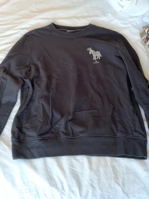 Mörkgrå sweatshirt med zebrapatch - Mörkgrå sweatshirt med rund hals och lång ärm. På bröstet finns en liten vit zebrapatch med texten 'Paul Smith'. Tröjan har en avslappnad passform och är tillverkad i mjukt bomullsmaterial.