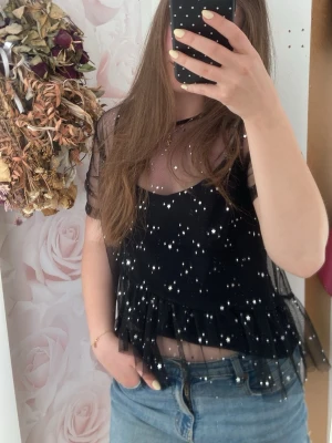 Svart mesh topp med glitterstjärnor - Superfin svart mesh topp med glittriga stjärnor och volang nertill. Toppen är genomskinlig och har korta ärmar, perfekt att styla över ett linne eller bandeau. Stjärnorna kan lätt ramla av❣️