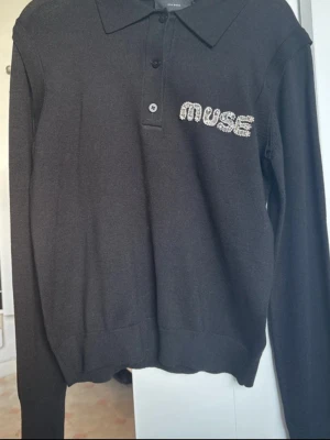 Svart tröja med strassdetalj från CPH Muse - Svart långärmad tröja från CPH Muse med krage och knäppning framtill. På bröstet står det 'MUSE' i glittrande strass. Tröjan har en klassisk passform och är perfekt för dig som gillar stilrena detaljer.