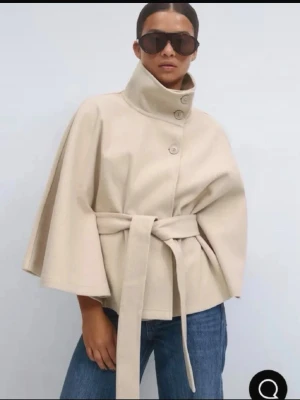 Beige cape-kappa med bälte - Stilren beige cape-kappa med hög krage, knäppning framtill och matchande knytskärp i midjan. Vida ärmar och avslappnad passform ger en trendig siluett. Perfekt att slänga över en hoodie eller stickad tröja för en cool look.