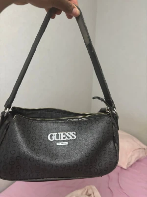   Väska  - En svart handväska från Guess. Man ser att den är lite rostig där man håller i väskan 