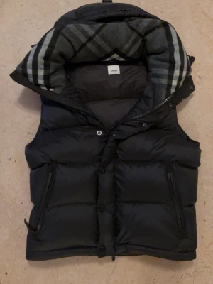 Svart dunväst från Burberry - Snygg svart dunväst från Burberry med klassiskt rutigt foder i huvan. Västen har dragkedja och knappar framtill samt två fickor med dragkedja. Perfekt för lager-på-lager och ger en stilren look med ikoniska Burberry-detaljer.
