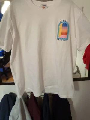 Vit Casa Blanca t-shirt med tryck - Vit t-shirt från Casa Blanca med färgglatt grafiskt tryck på bröstet och ryggen. Trycket föreställer en bågformad öppning med gradientfärger i blått, gult, rosa och orange. Klassisk passform och rund hals. Perfekt för en chill och trendig look.