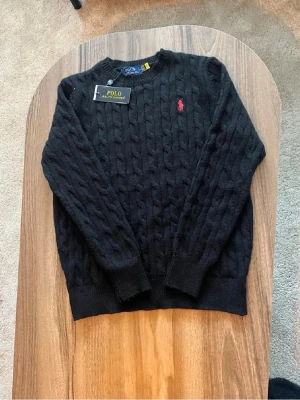 Svart kabelstickad tröja Polo Ralph Lauren - Kabelstickad svart tröja från Polo Ralph Lauren med rund halsringning och den klassiska röda logotypen broderad på bröstet. Tröjan har ribbade muddar vid ärmslut och nederkant. Perfekt för dig som gillar stilrena och tidlösa plagg.