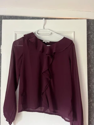 Vinröd blus med volang från SAMSA - Vinröd blus från SAMSA YOUNG i storlek 146/152. Blusen har långa ärmar och en snygg volangdetalj längs halsringningen och framtill. Perfekt för dig som gillar en feminin och trendig stil. I mycket bra skick❣️