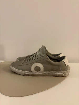  Oliver Cabell sneakers  - Snygga gråa sneakers från Oliver Cabell med vit häl och vit cirkeldetalj på sidan. Skorna har klassisk låg profil, snörning och är tillverkade i läder. Perfekta för en clean och stilren look. Kvitto finns!