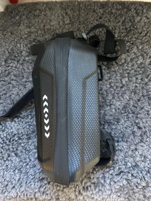 Svart hårdskalad elscooter/cykelväska - Säljer en svart hårdskalad väska med hexagonmönster och vita pilar på sidan. Väskan har justerbara remmar och är tillverkad i slitstarkt syntetmaterial. Perfekt för att fästa på cykeln eller elkicken man kan också använda den som en liten förvaringsväska.