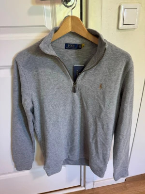 Grå halvzip från Polo Ralph Lauren - Snygg grå långärmad tröja från Polo Ralph Lauren med halv dragkedja och klassisk broderad logga på bröstet. Tröjan har en hög krage och är tillverkad i mjukt material som passar perfekt till lager-på-lager-stil.