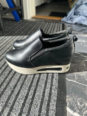 Svarta sneakers med platåsula - Säljer ett par svarta sneakers med hög vit platåsula och slip-on design. Skorna har en modern look med släta ytor och elastiska paneler på sidorna för enkel påtagning. Materialet är syntetiskt och sulan har coola detaljer. Perfekt för dig som gillar att sticka ut.